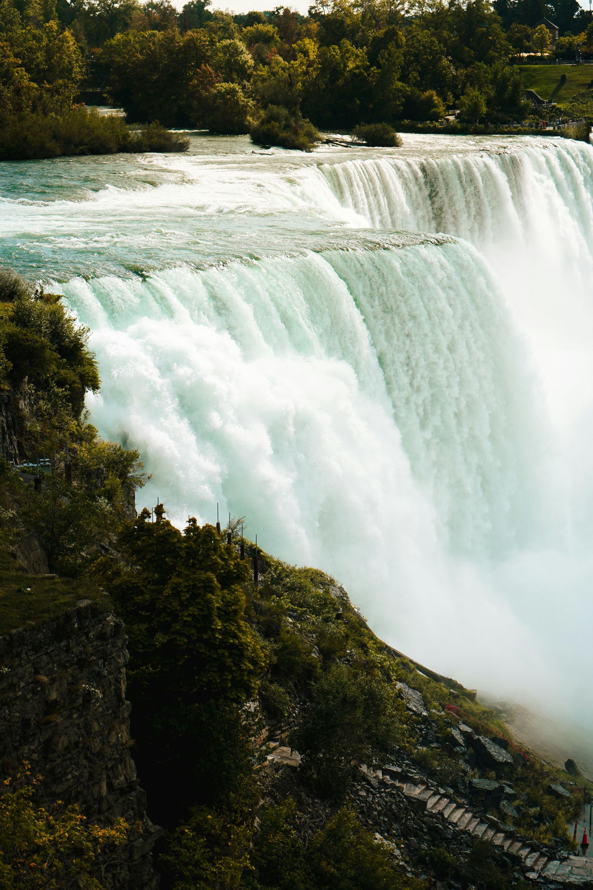 A powerful, wide waterfall cascades over a rocky cliff surrounded by green trees, creating misty spray at the bottom.