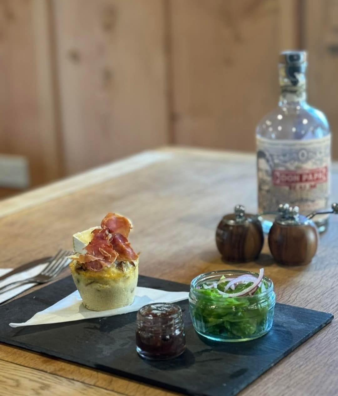 A quiche topped with bacon, a small jar of relish, and a side salad on a slate board, with a rum bottle in the background.