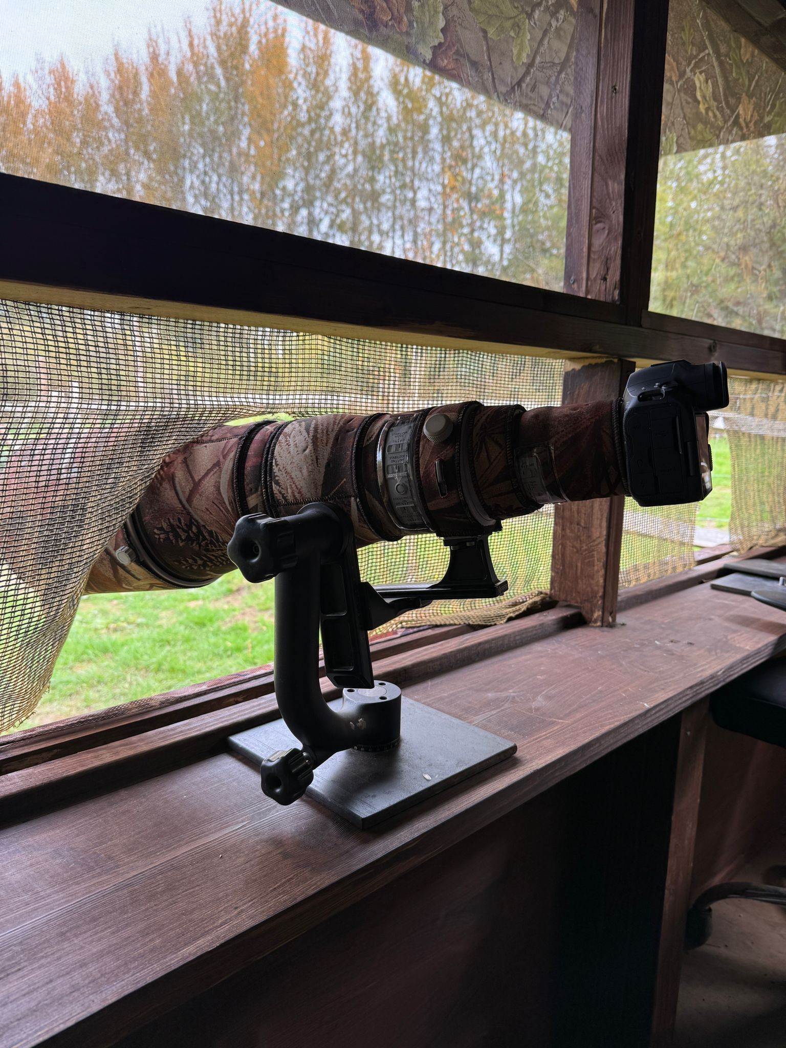 A camera with a camouflage lens cover mounted on a gimbal head, positioned on a wooden ledge inside a wildlife hide.