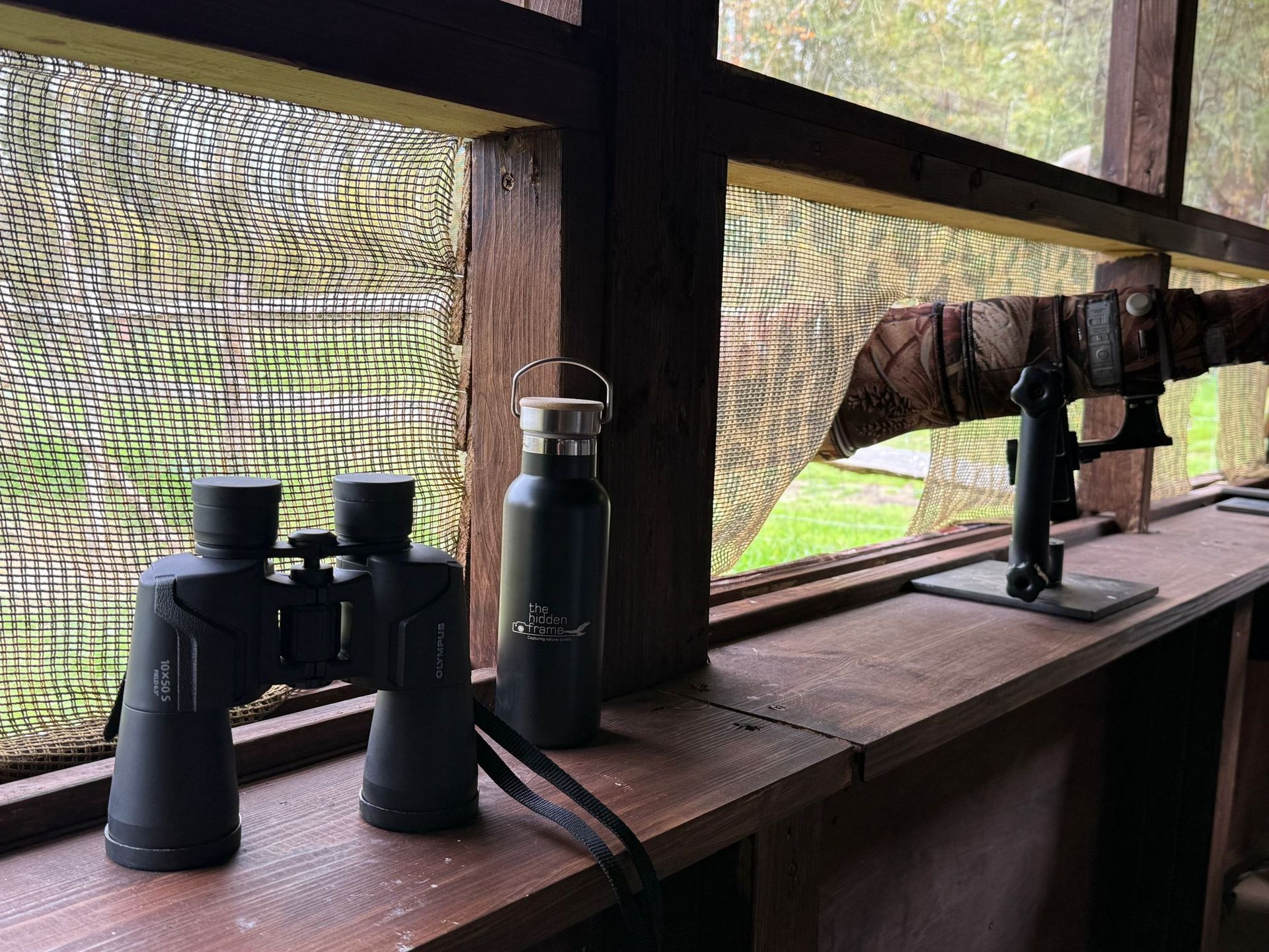 Binoculars and a metal water bottle sit on a wooden ledge inside a camouflaged hunting blind.