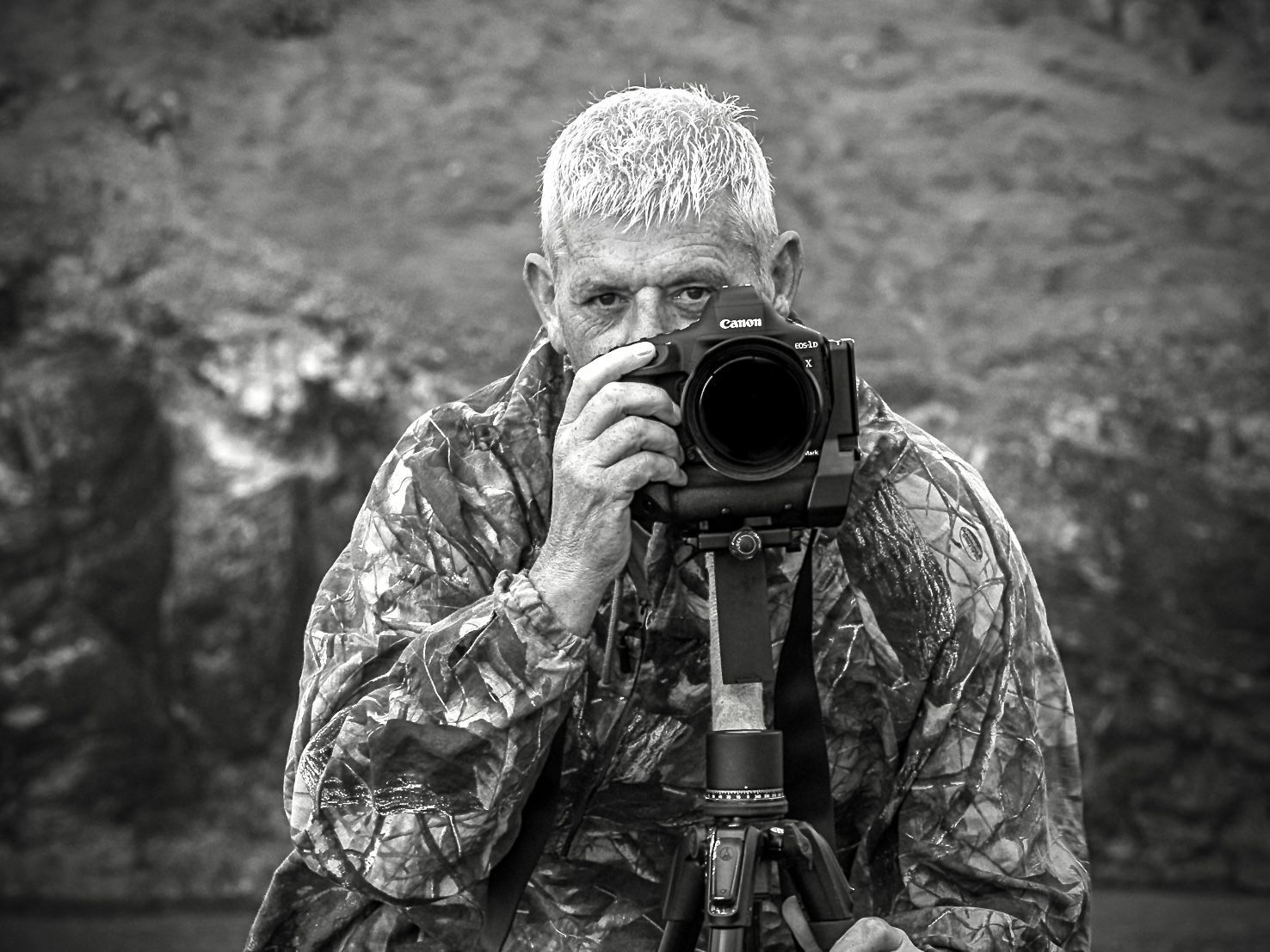 A person in a camouflage jacket behind a camera mounted on a tripod, appearing to photograph the viewer.