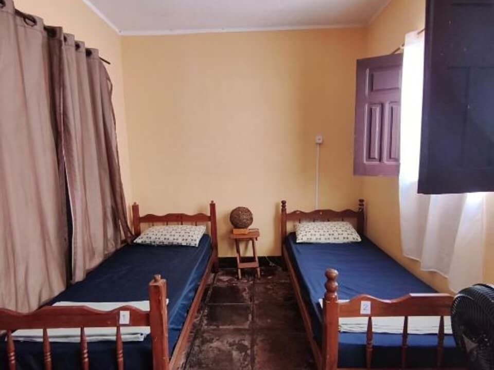 Quarto pequeno com duas camas de solteiro, cortinas e uma janela com entrada de luz.