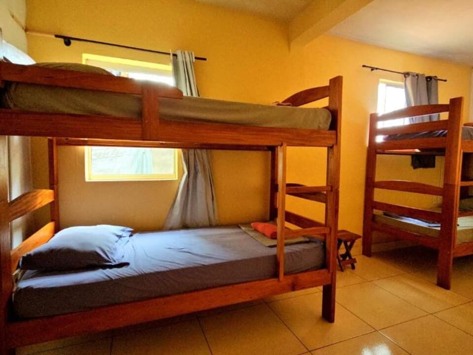 Quarto de dormitório com beliches de madeira, roupa de cama cinza, paredes amarelas e janelas com cortinas.