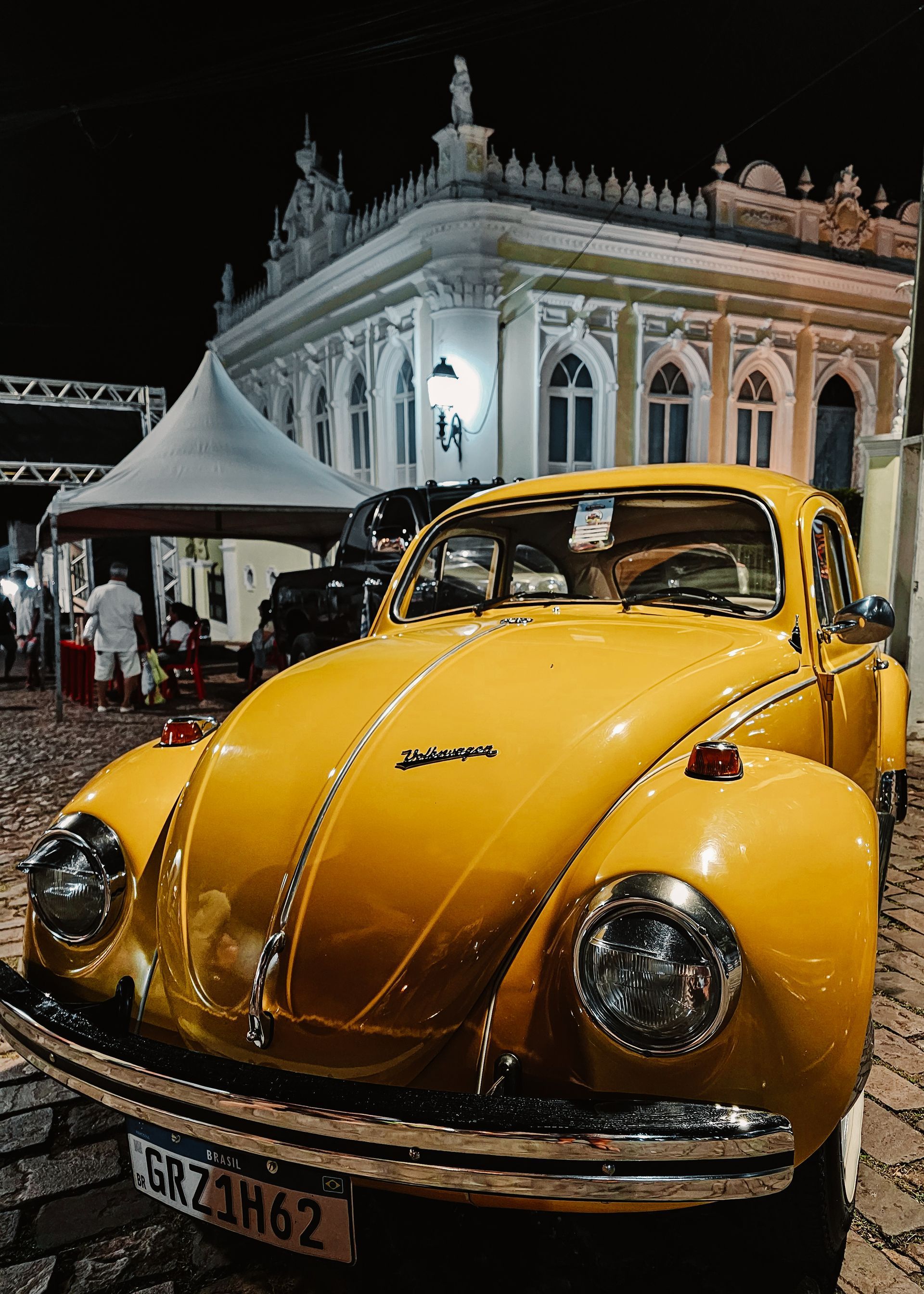 Fusca Volkswagen amarelo antigo estacionado à noite em frente a um prédio branco ornamentado.