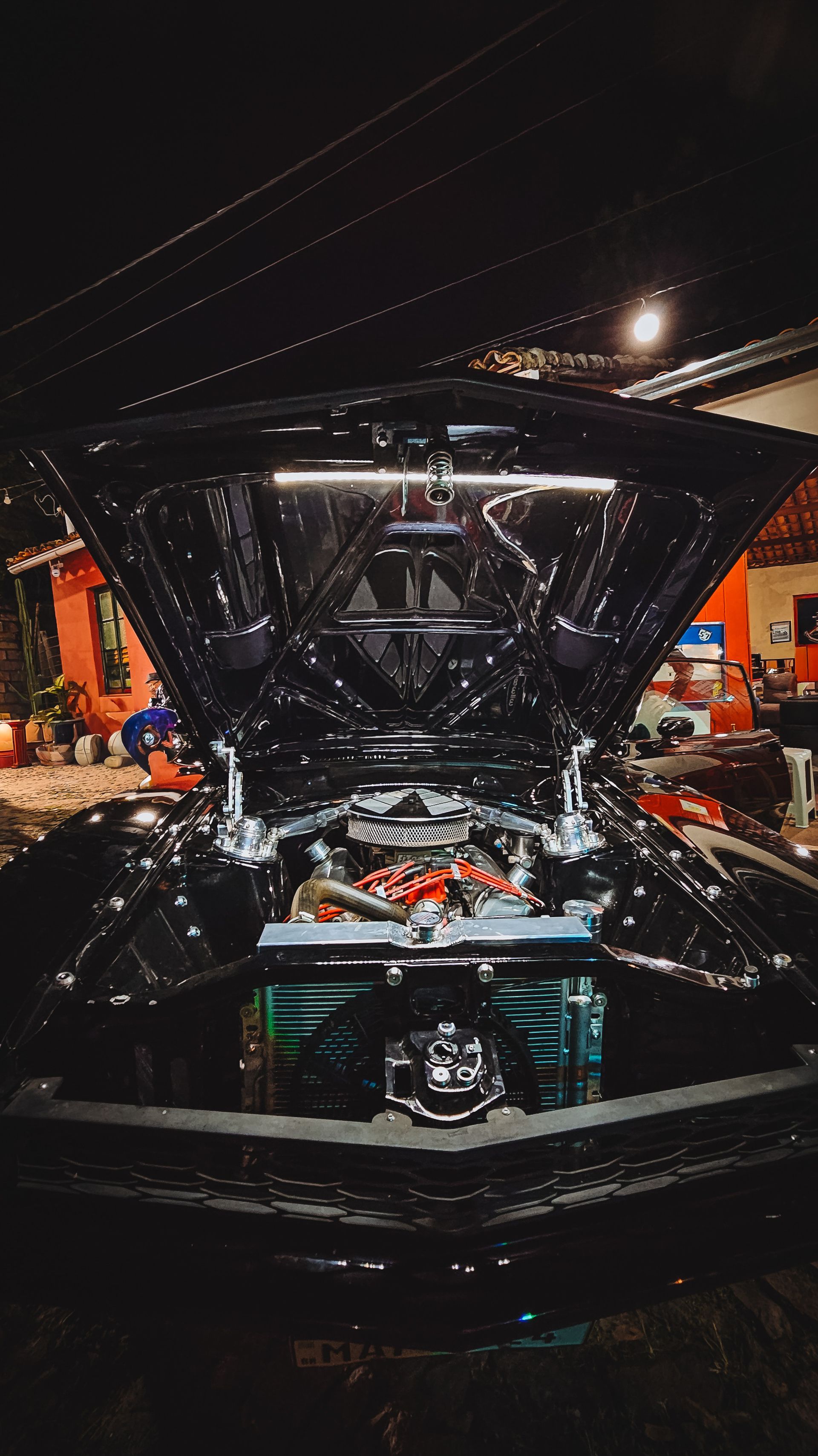 Capô aberto de um carro com o compartimento do motor exposto em uma exposição de automóveis ou garagem à noite.
