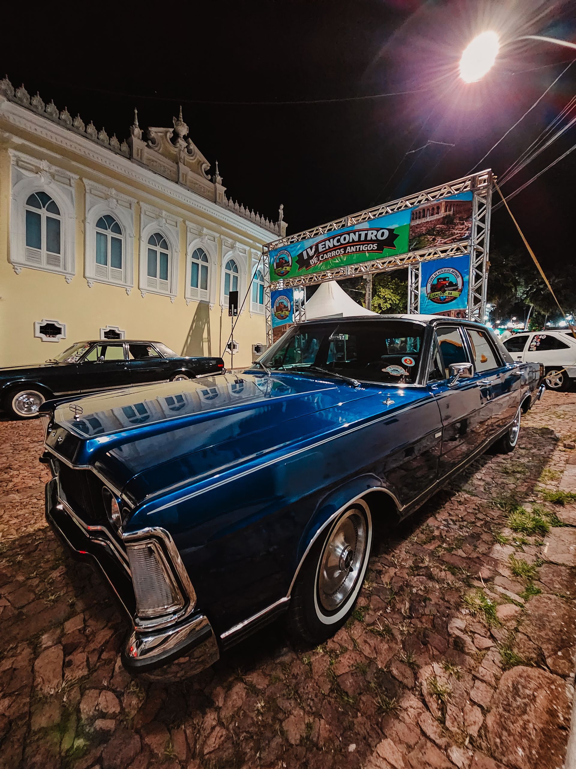 Carro antigo azul em uma rua de paralelepípedos à noite, ao lado de um prédio iluminado e um palco para eventos.