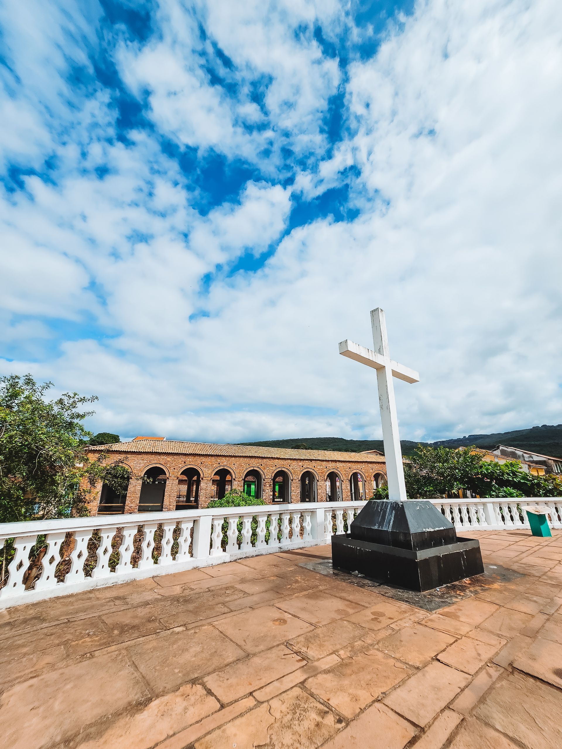 Cruz branca em um terraço de pedra com vista para uma longa estrutura arqueada sob um céu claro e nublado.