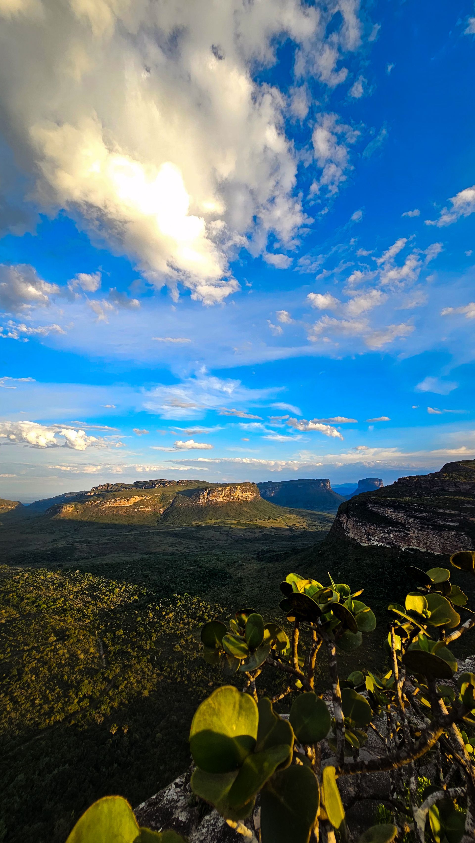 Crista de montanha iluminada pelo sol sob um céu azul com nuvens, com vista para um vale verde ao pôr do sol.