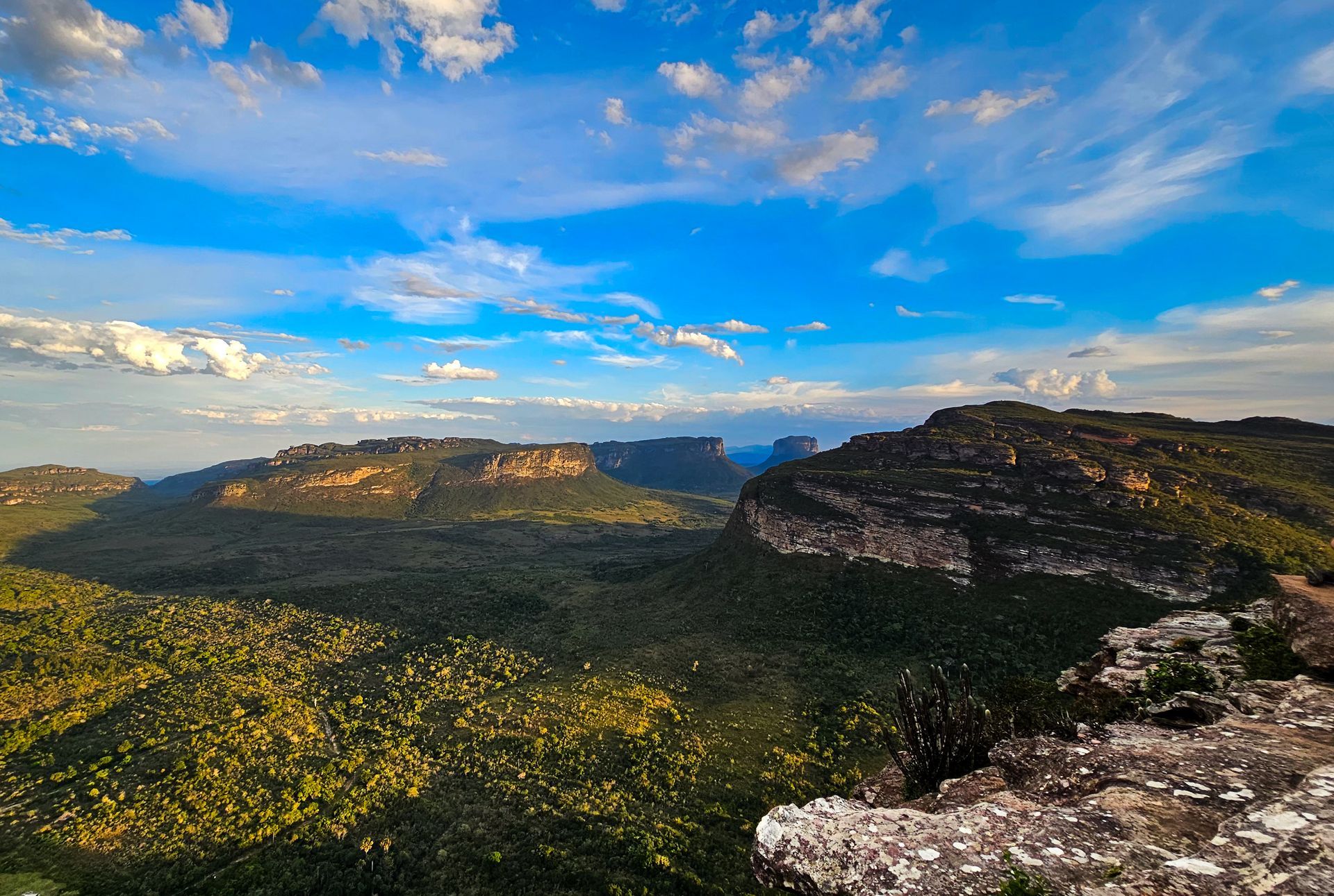Amplo vale montanhoso com florestas verdejantes, penhascos rochosos e um céu azul brilhante com nuvens dispersas.