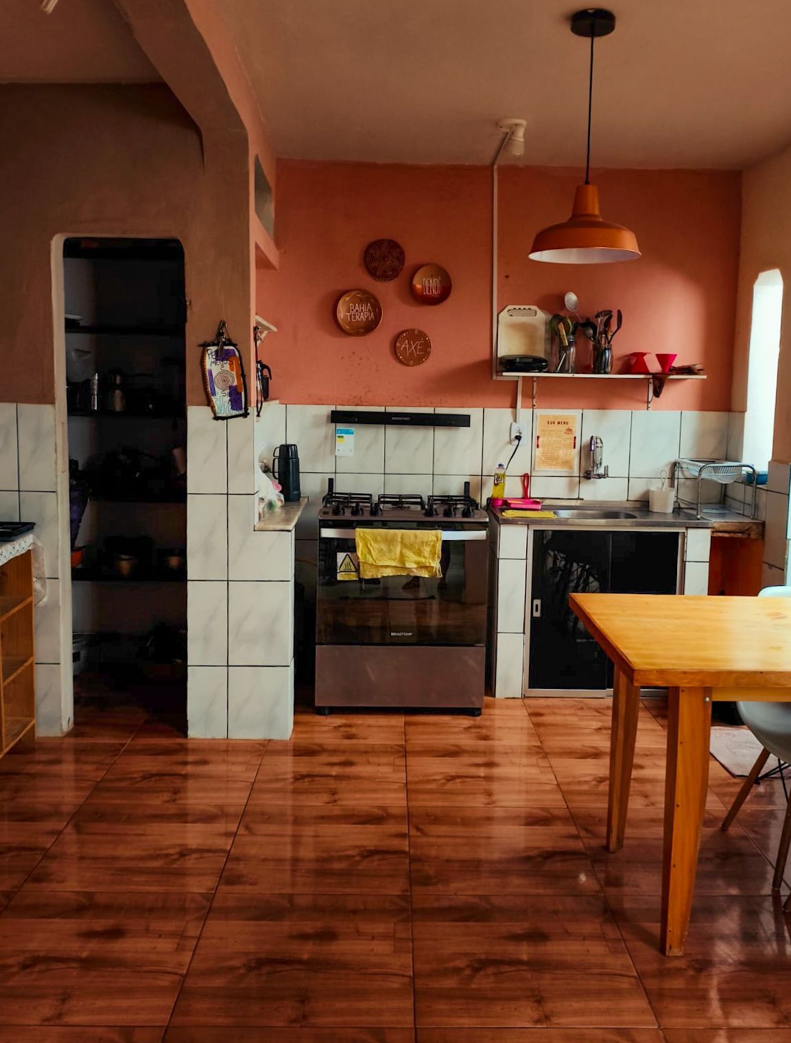Cozinha aconchegante com paredes laranja, piso de azulejo, fogão, armários e uma mesa de madeira junto a uma janela iluminada.