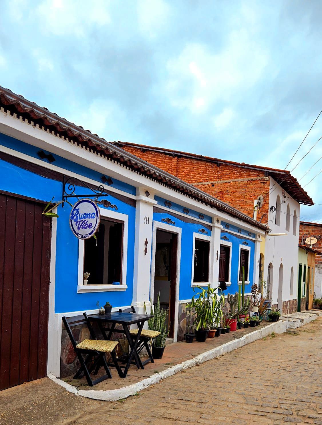 Café azul vibrante em rua de paralelepípedos, com mesas ao ar livre e telhado de telhas vermelhas sob um céu nublado.