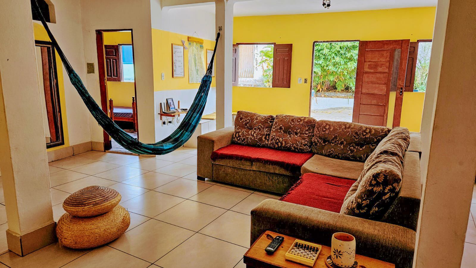 Sala de estar iluminada com uma rede, azulejos bege, sofás vermelhos e paredes amarelas banhadas pela luz do dia.