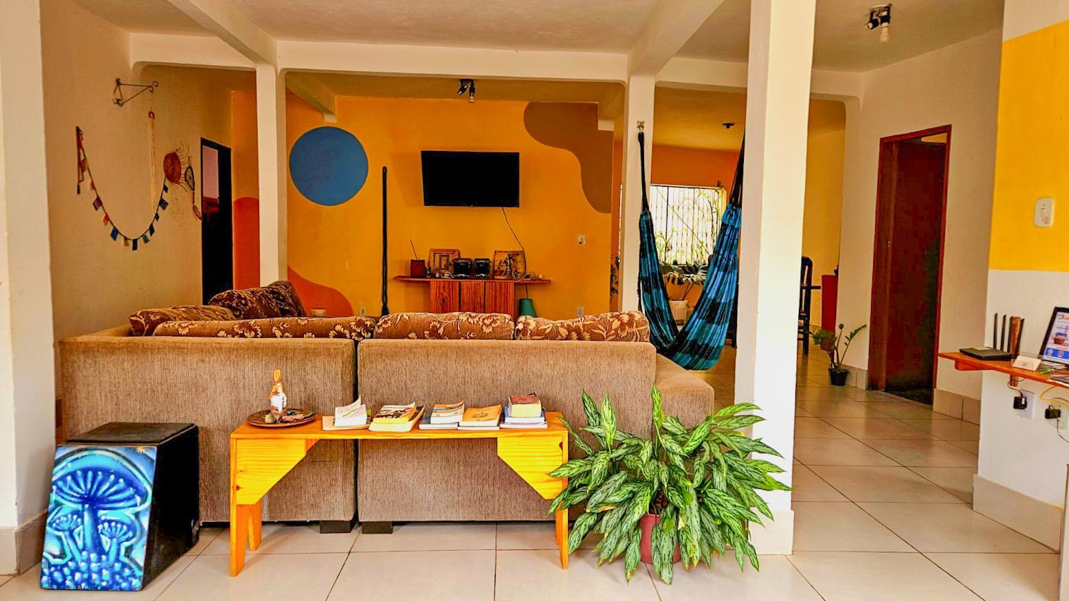 Sala de estar aconchegante com paredes laranja, sofá, TV, mesa, plantas e piso de azulejo branco.
