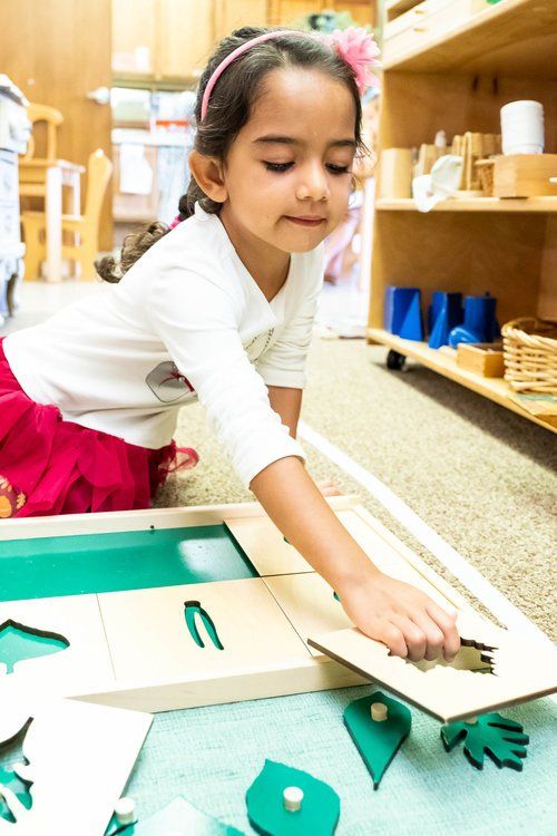 Child reaching into a wooden puzzle board in a classroom, with green and white pieces on a table
