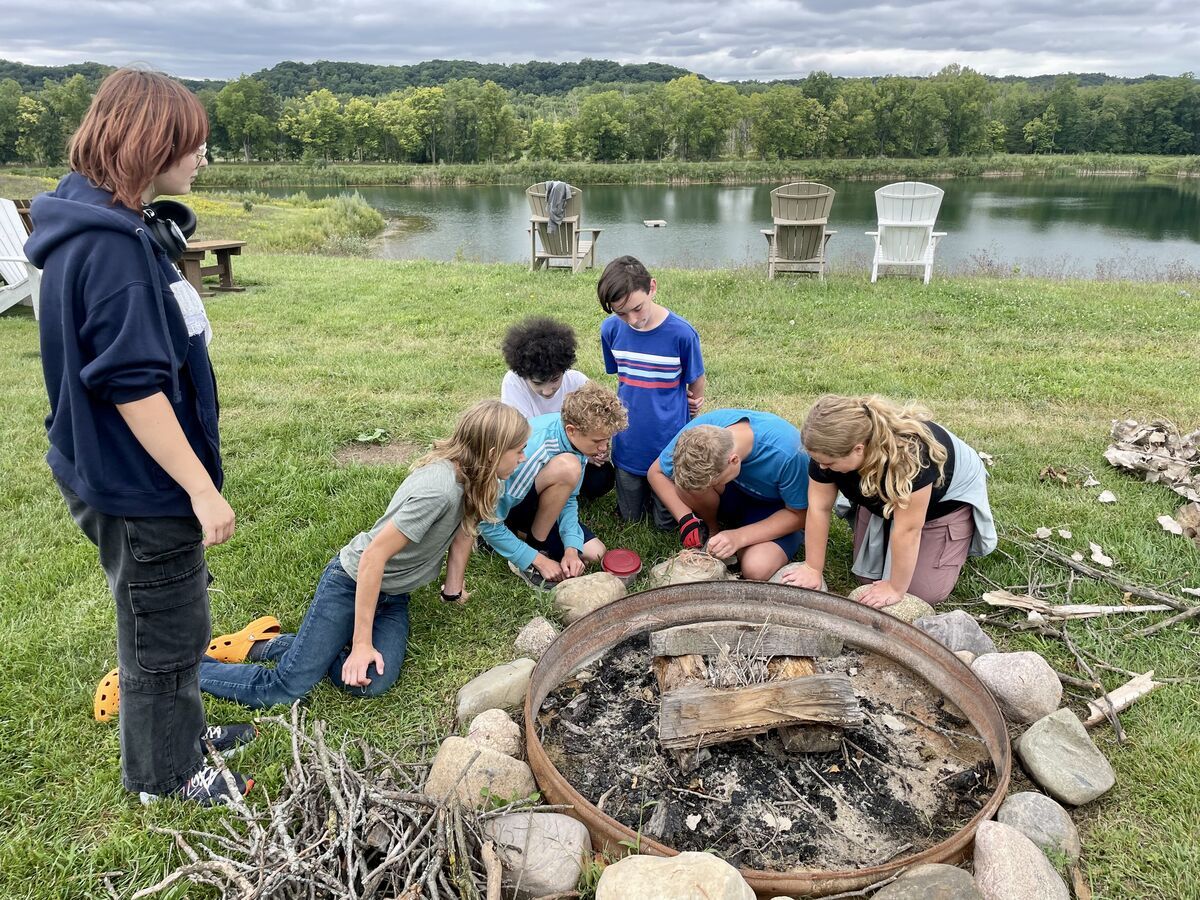 montessori children gathered around a stone fire pit by a pond in a grassy field, examining something inside.