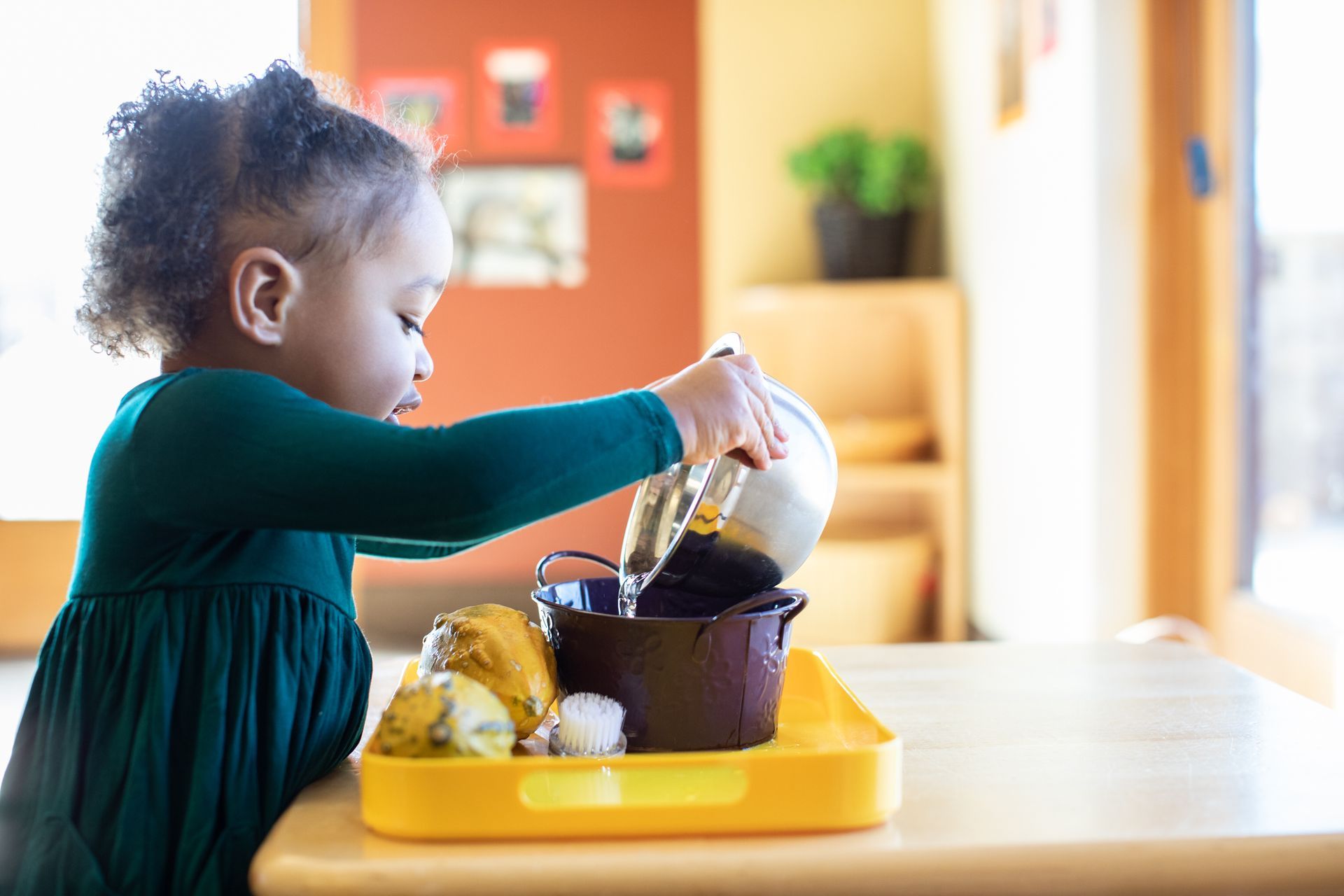 Child pouring from a glass pitcher into a bowl on a yellow tray at a table indoors