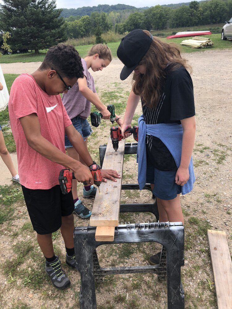 Montessori children sawing a wooden plank on a metal stand outdoors at a campsite