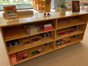 Montessori wooden classroom shelf with colorful books and Montessori materials beneath a window