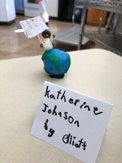 Small handmade figure with a blue-green body and white head, labeled “Katherine Johnson by Eliott” on a card.