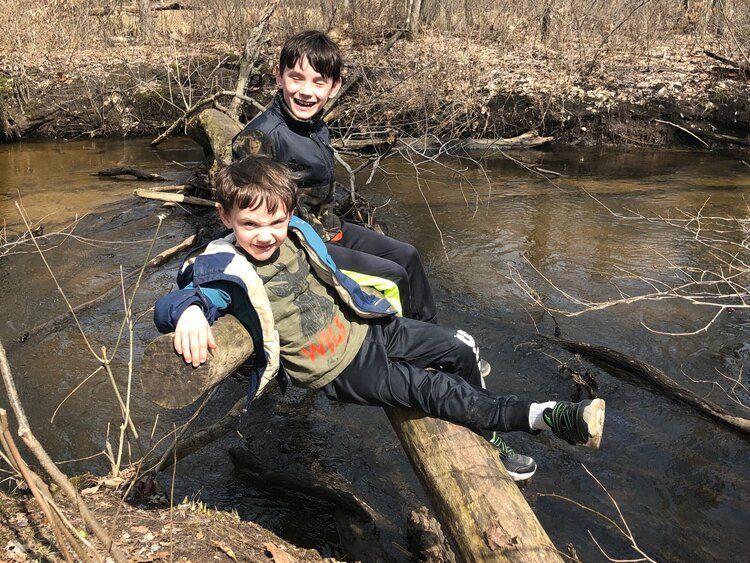 Two montessori children smiling while balancing on a fallen tree over a shallow creek in a wooded area.