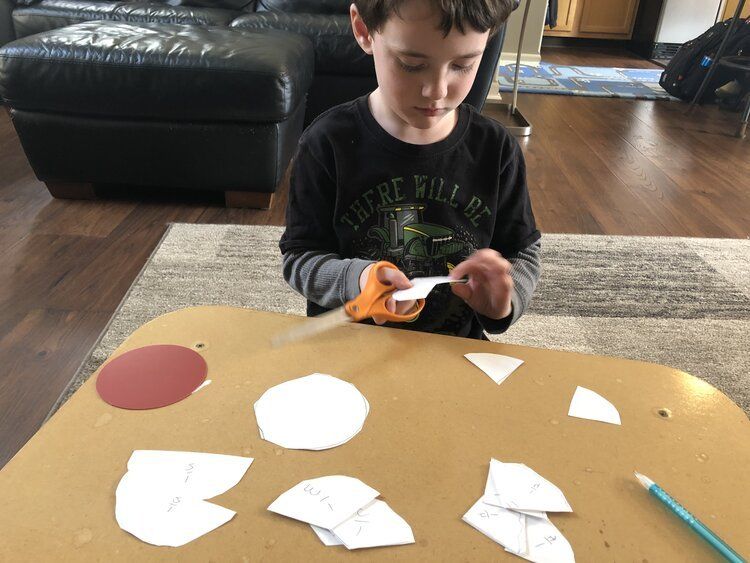 Montessori child cutting white paper shapes at a table with scissors and a blue pencil nearby.