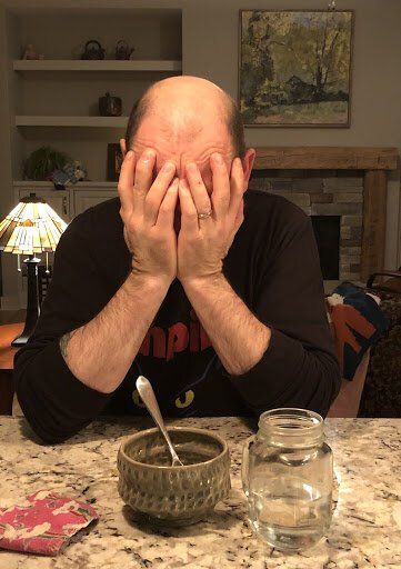 Man sitting at a kitchen table with hands covering his face, with a bowl and jar in front of him.