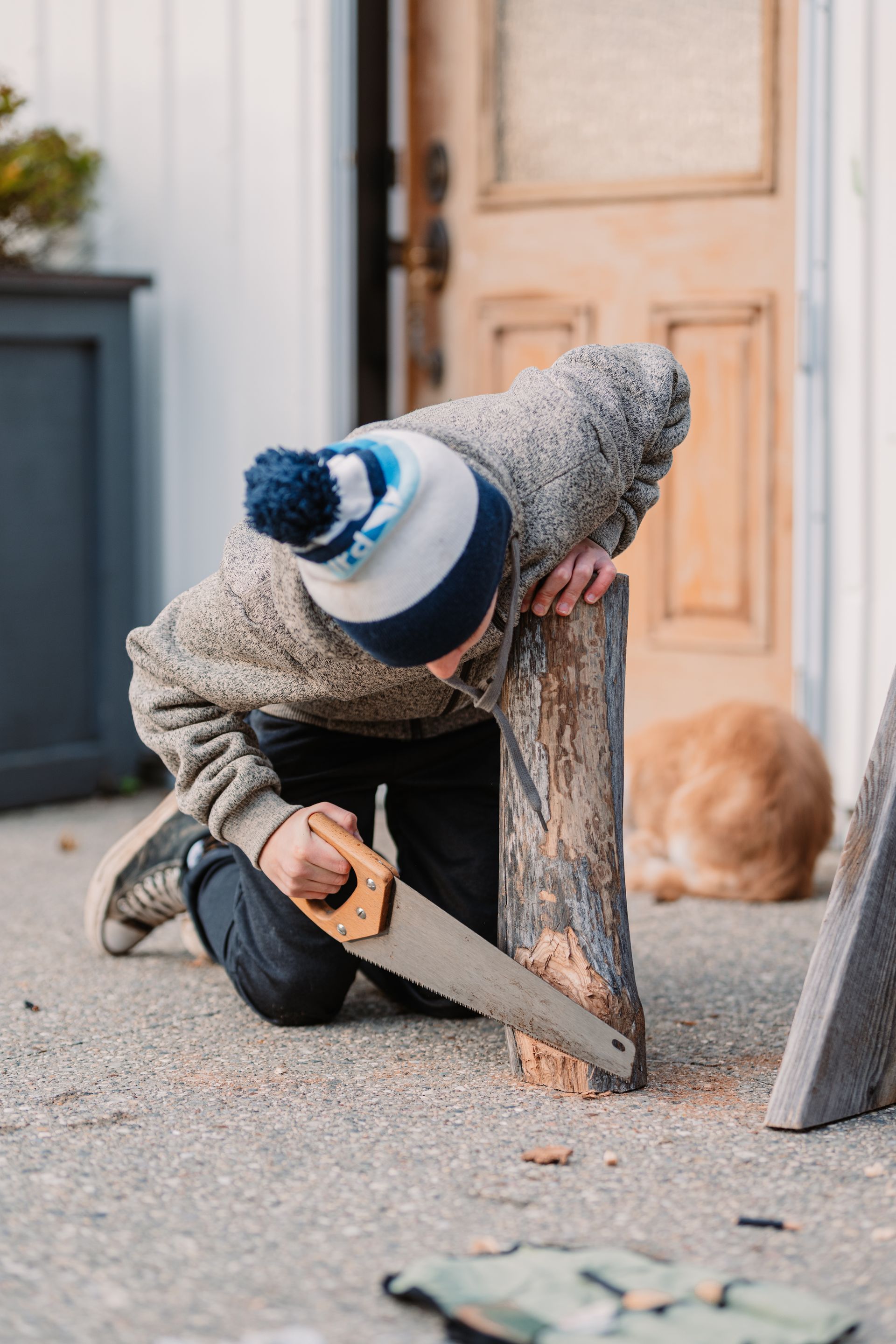 Person cutting a wooden post with a hand saw outside a door, wearing a striped knit hat and sweater.