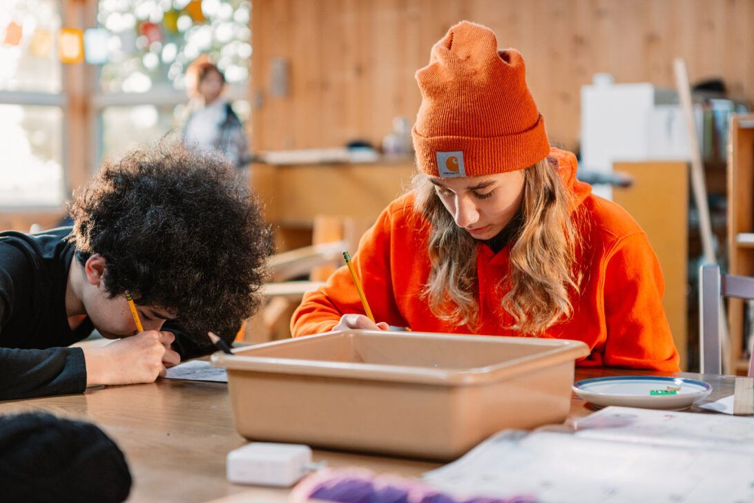 Two montessori children drawing at a table, with one in an orange beanie and sweater in a classroom.