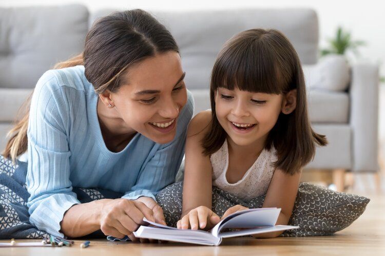 Woman and child smiling while reading a book together on a living room floor