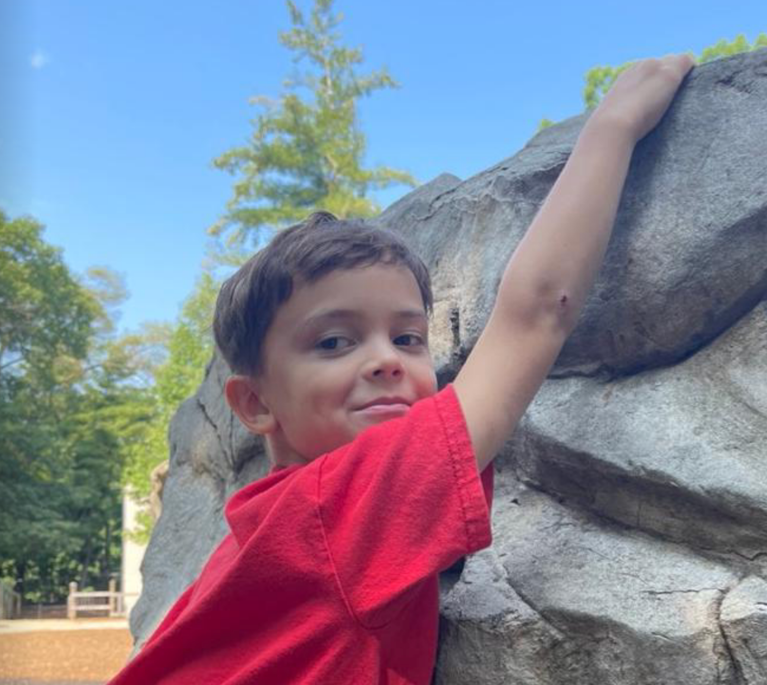 Montessori child in a red shirt climbing a large rock outdoors, smiling at the camera.