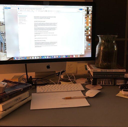 Desk with iMac displaying a document, keyboard, papers, cables, and a glass pitcher beside books.