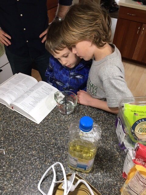 Two Montessori children reading a book together at a kitchen counter beside snacks and a bottle of oil
