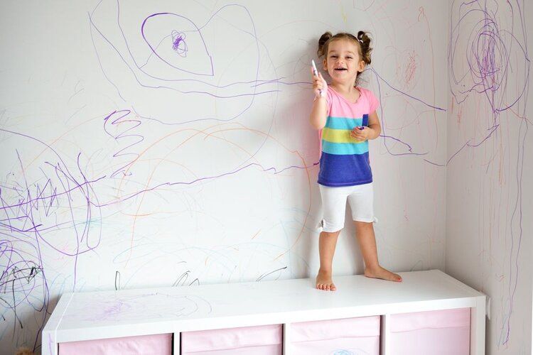 Child standing on a white cabinet with purple crayon drawings on the wall behind her