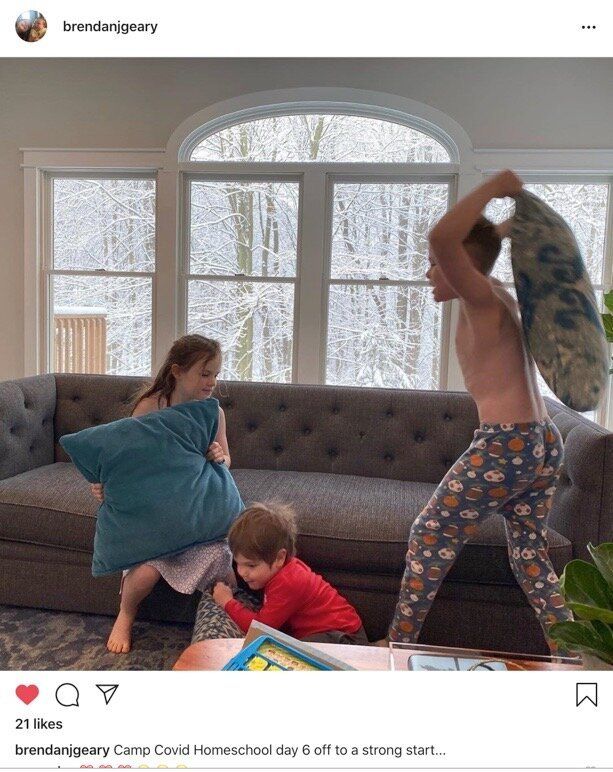 Two montessori children play on a living room floor while an adult swings a blue pillow nearby.