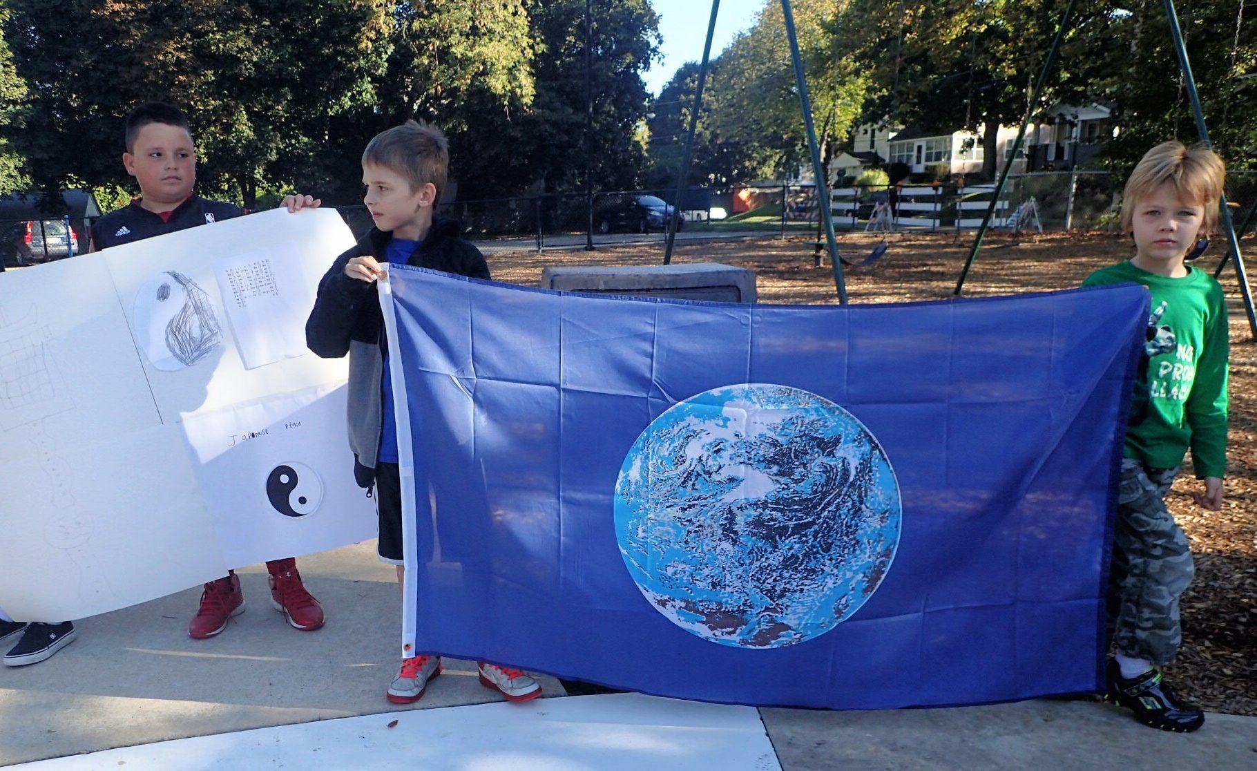 Three Montessori children holding white and blue flags outdoors in a sunny park
