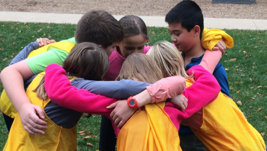 Montessori children in colorful tops huddle in a circle with arms around each other outdoors