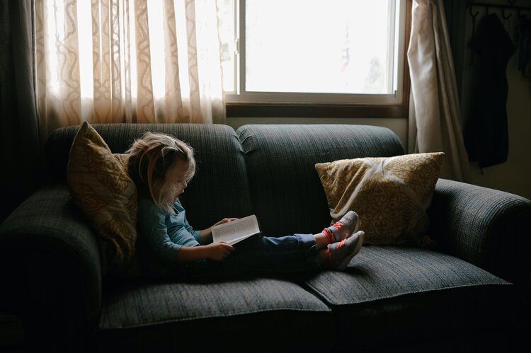 Montessori Child lying on a dark couch reading a book by a sunlit window, with pillows nearby.