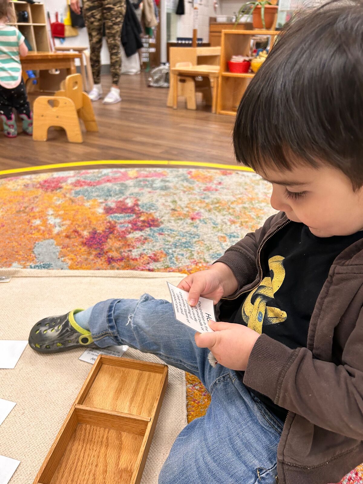 montessori Child sitting on the floor, reading a card beside a wooden tray in a colorful playroom.