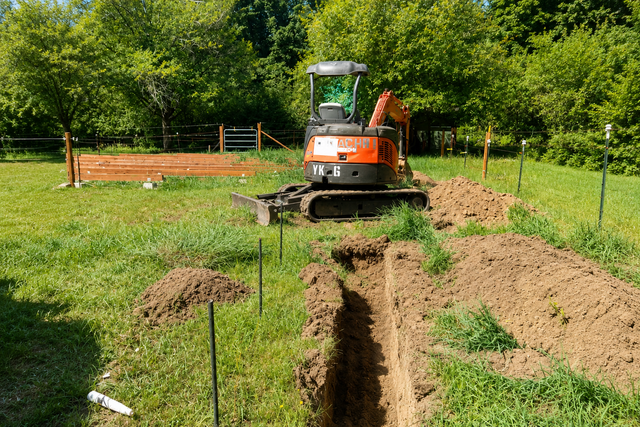 Mini excavator digging a trench in a grassy yard with soil piles and fencing.