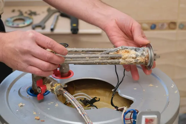 Hands remove a corroded heating element from a water heater tank.