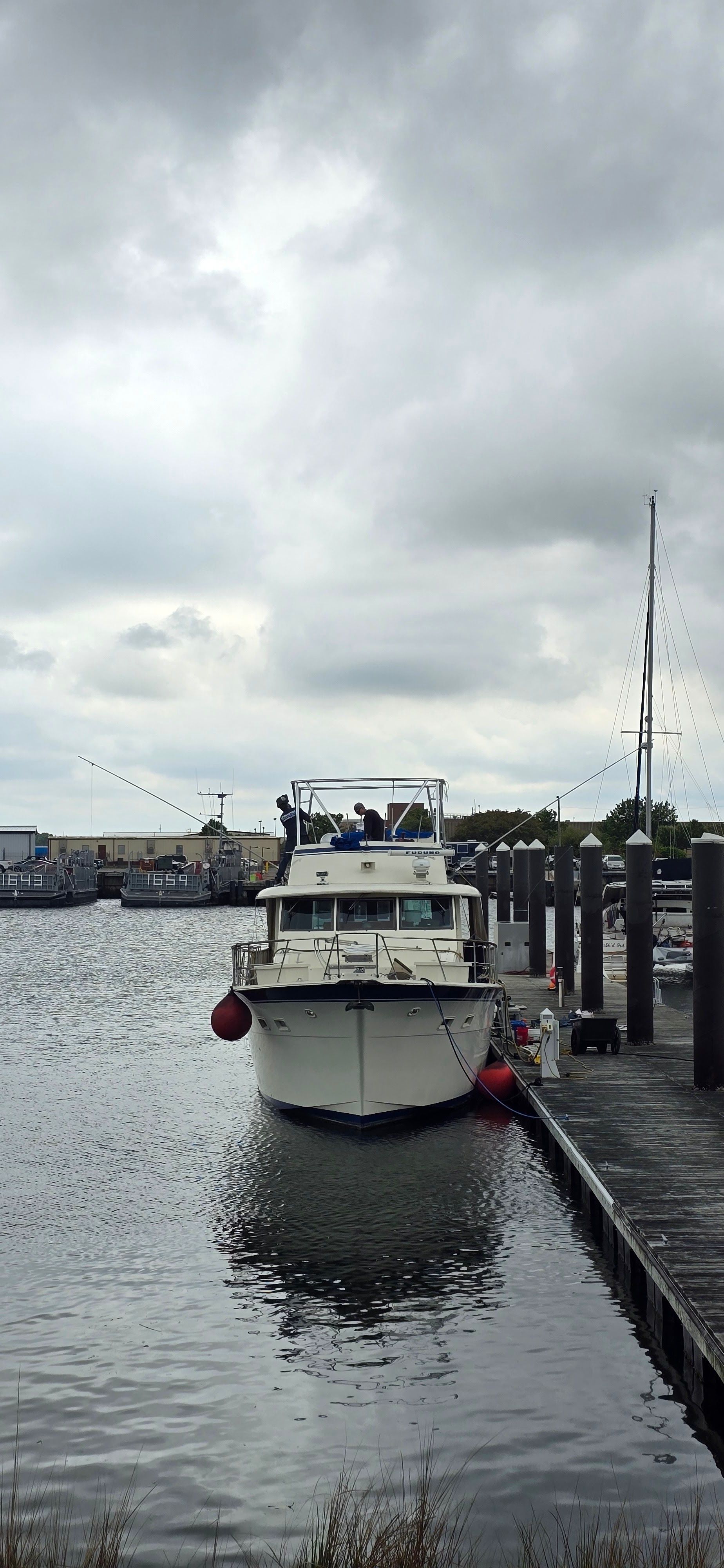 White boat docked in a narrow harbor under cloudy skies, with water and moored posts alongside.