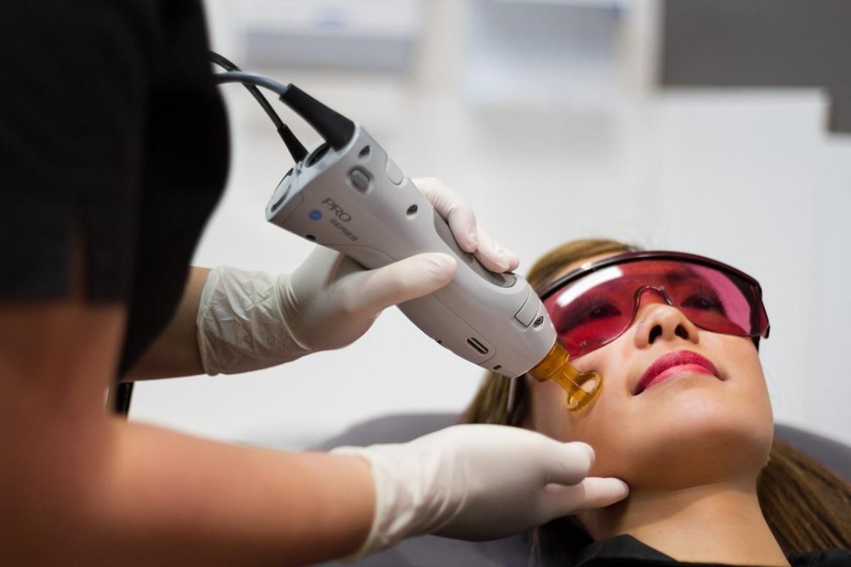 Person receiving facial laser treatment, wearing safety glasses. Doctor holds device to their cheek in a medical setting.