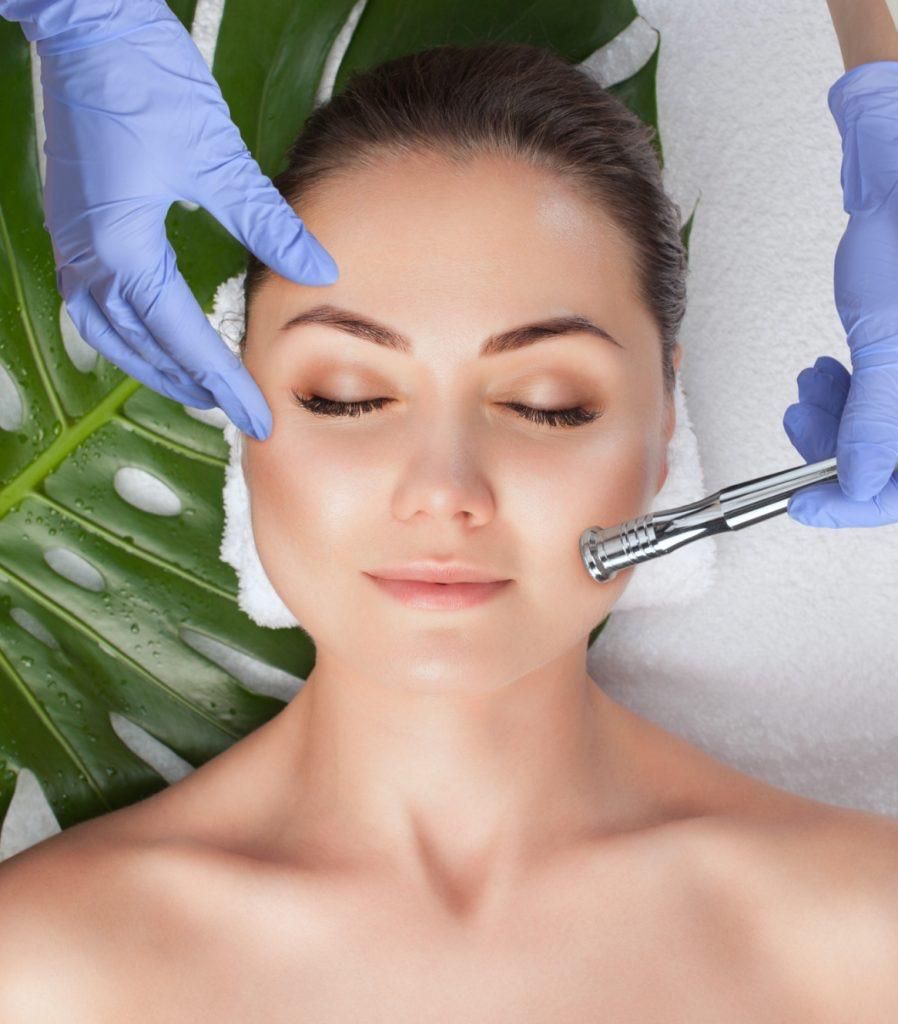 Woman receiving facial treatment, hands in blue gloves hold a tool near her face. Green leaf and white towel background.