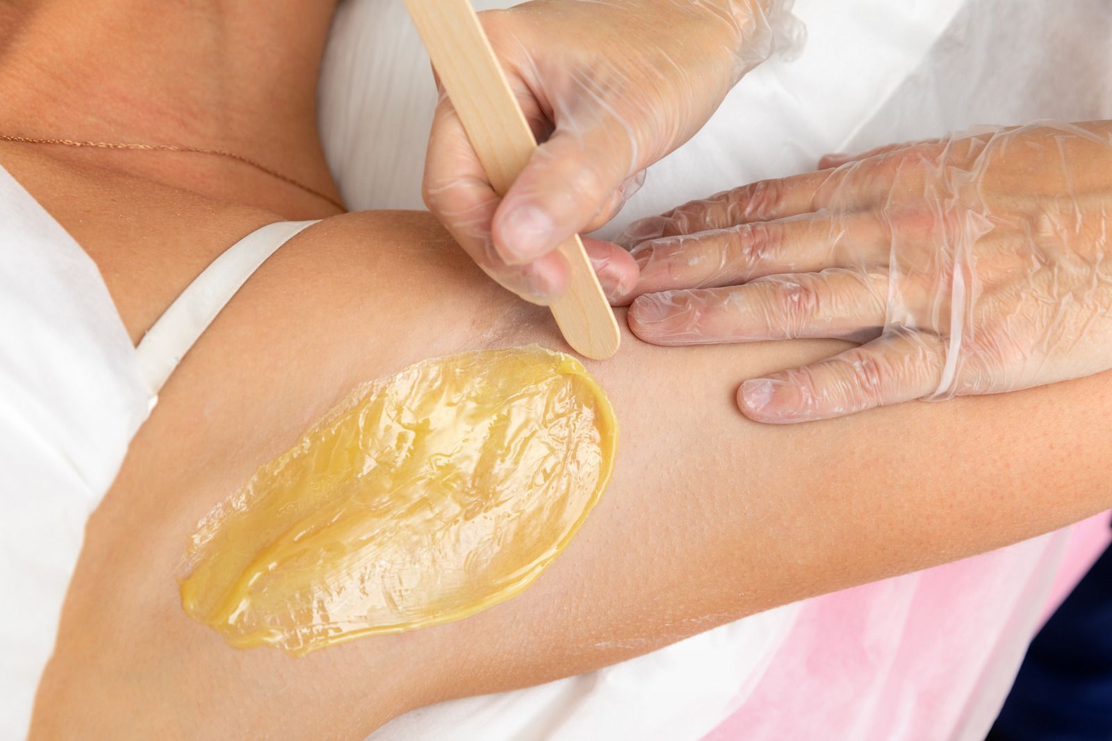 Wax being applied to underarm with gloved hands using a wooden stick.