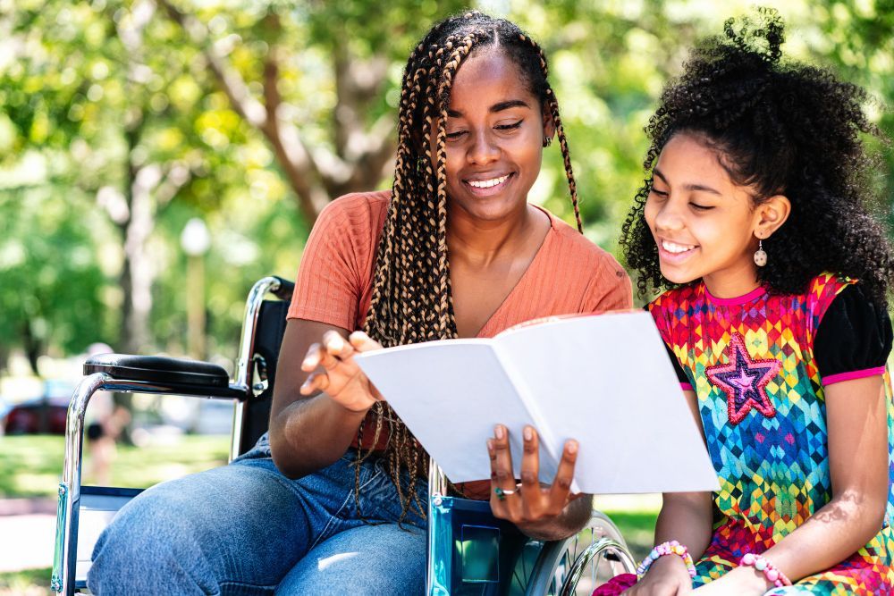 Two people smiling and reading a book together outdoors near a wheelchair