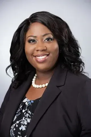Studio headshot of a smiling professional in a black blazer and pearl necklace on a light gray background