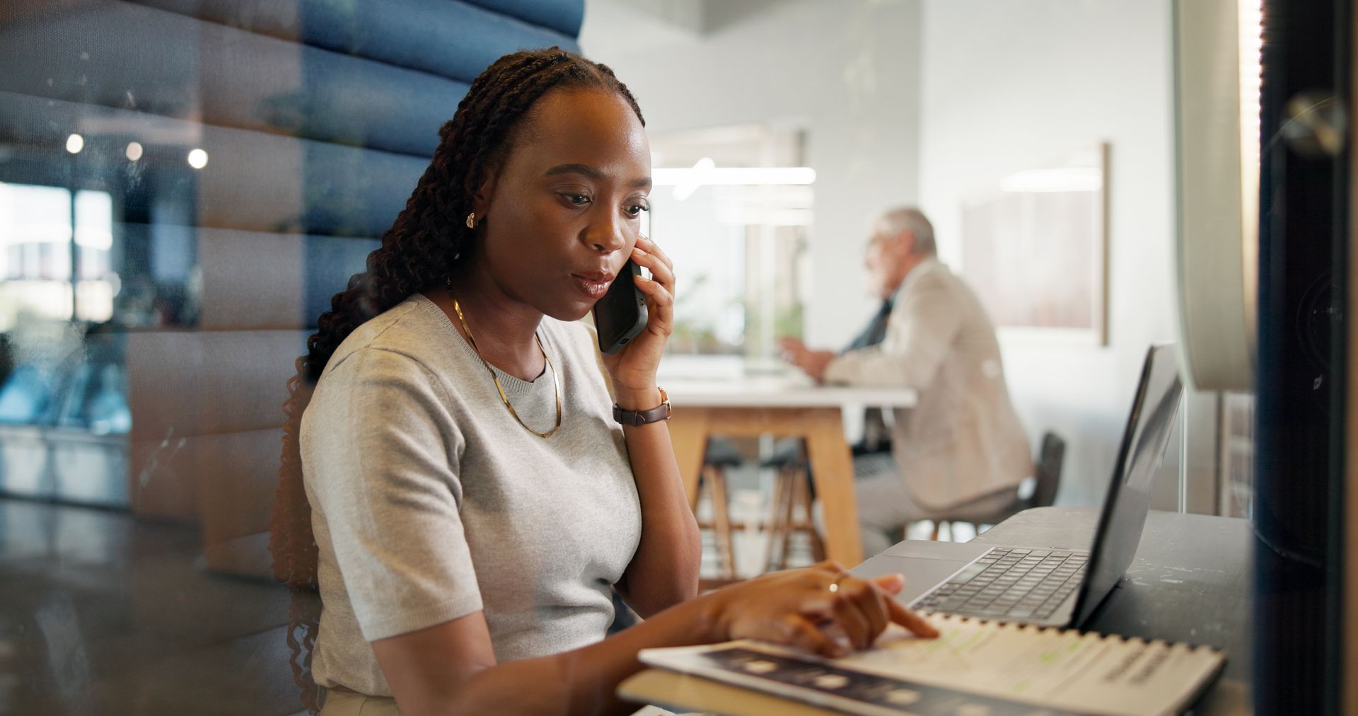 Woman working on a laptop at a table in a modern office with a blurred colleague in the background