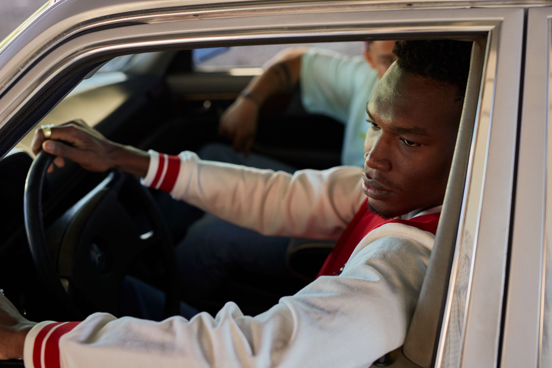 Man in a red-and-white jacket driving a car, looking out the open window with a focused expression
