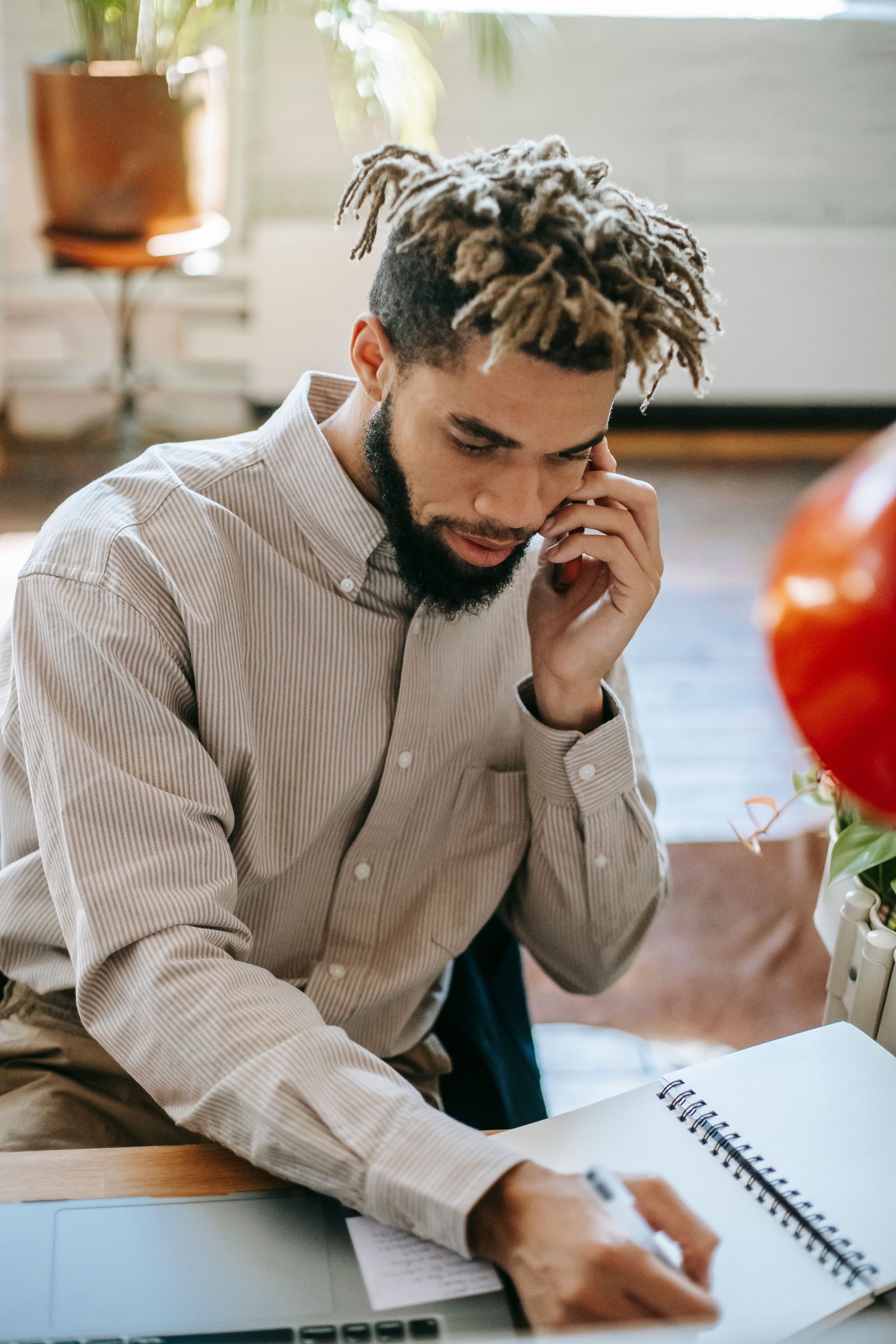 Man at a desk on the phone, writing in a notebook in a bright office.