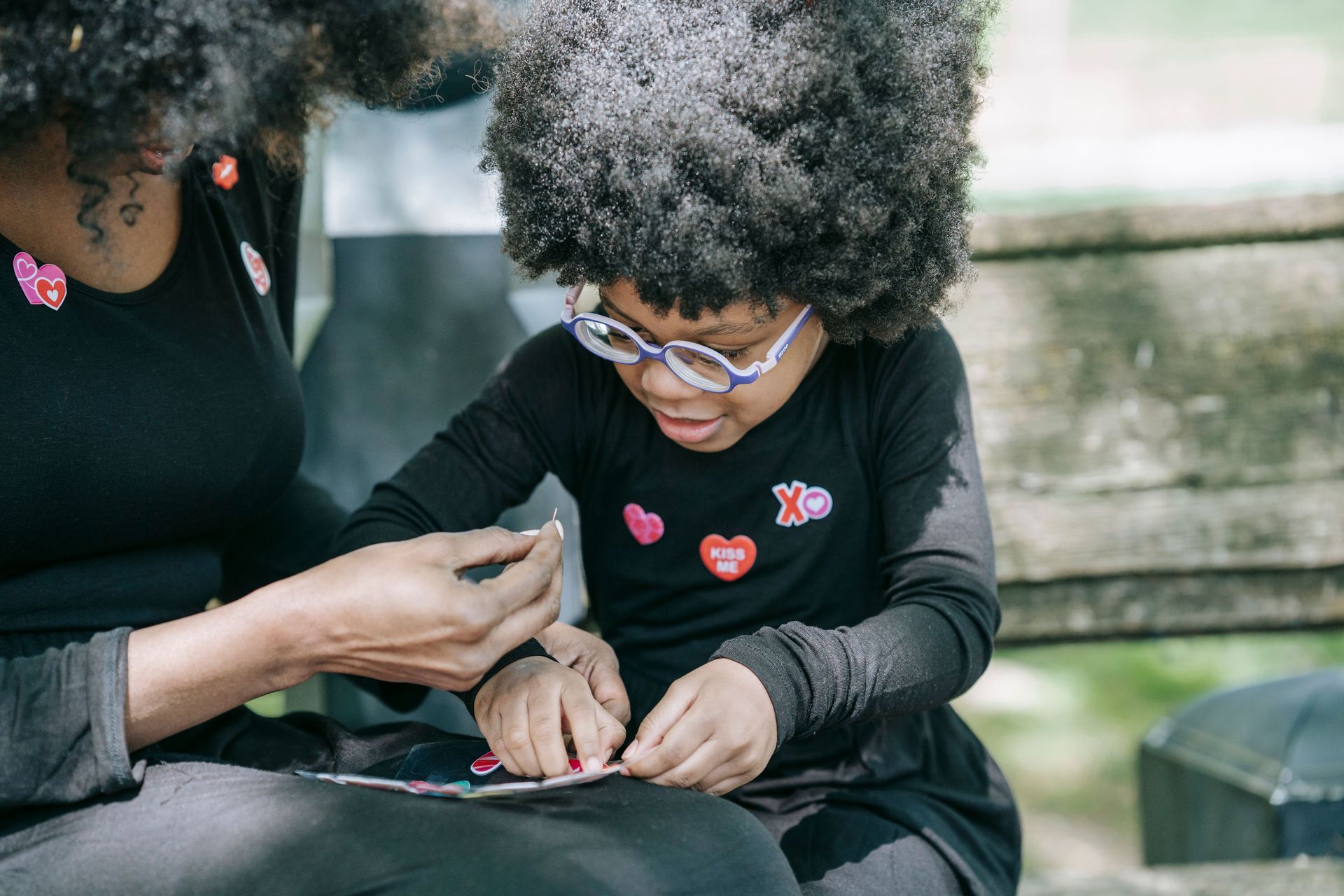 Adult helping child with hands, both in black shirts with heart patches, seated outdoors on a bench.
